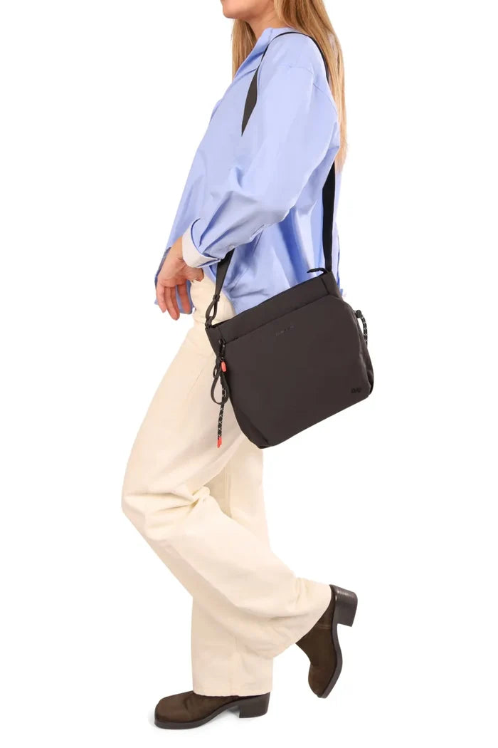 Hedgren MAKOTO Square Crossover Bag in Black, worn crossbody at the hip by a woman walking in a blue shirt and cream trousers