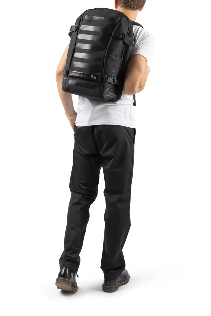 Hedgren TRIP M Expandable 15.6" Laptop Travel Backpack with RFID in Black, worn on both shoulders by a man in a white tee, seen from behind