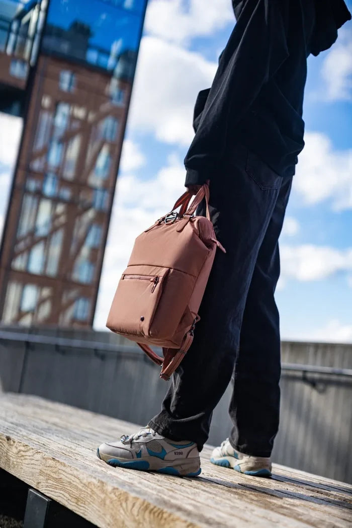 Pacsafe Citysafe CX Econyl Anti-Theft Mini Backpack in Rose, held by its top handles beside a woman standing on a bench