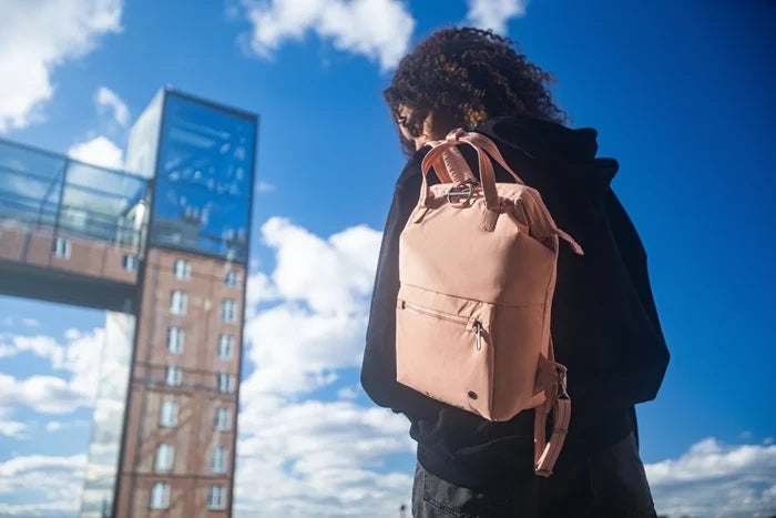 Pacsafe Citysafe CX Econyl Anti-Theft Mini Backpack in Rose, worn on both shoulders by a woman standing below a glass tower