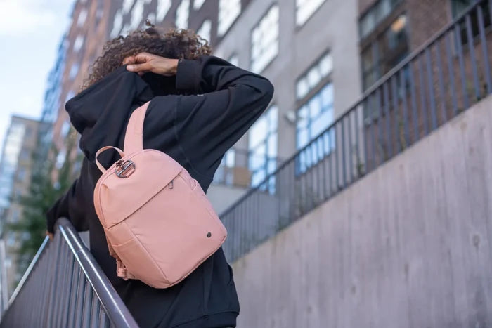 Pacsafe Citysafe CX Petite Anti-Theft 8L Backpack in Rose, swung off one shoulder by a woman on a city street