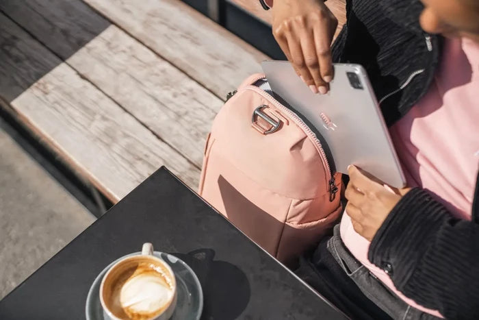 Pacsafe Citysafe CX Petite Anti-Theft 8L Backpack in Rose, a tablet being lifted out at a cafe table beside a coffee