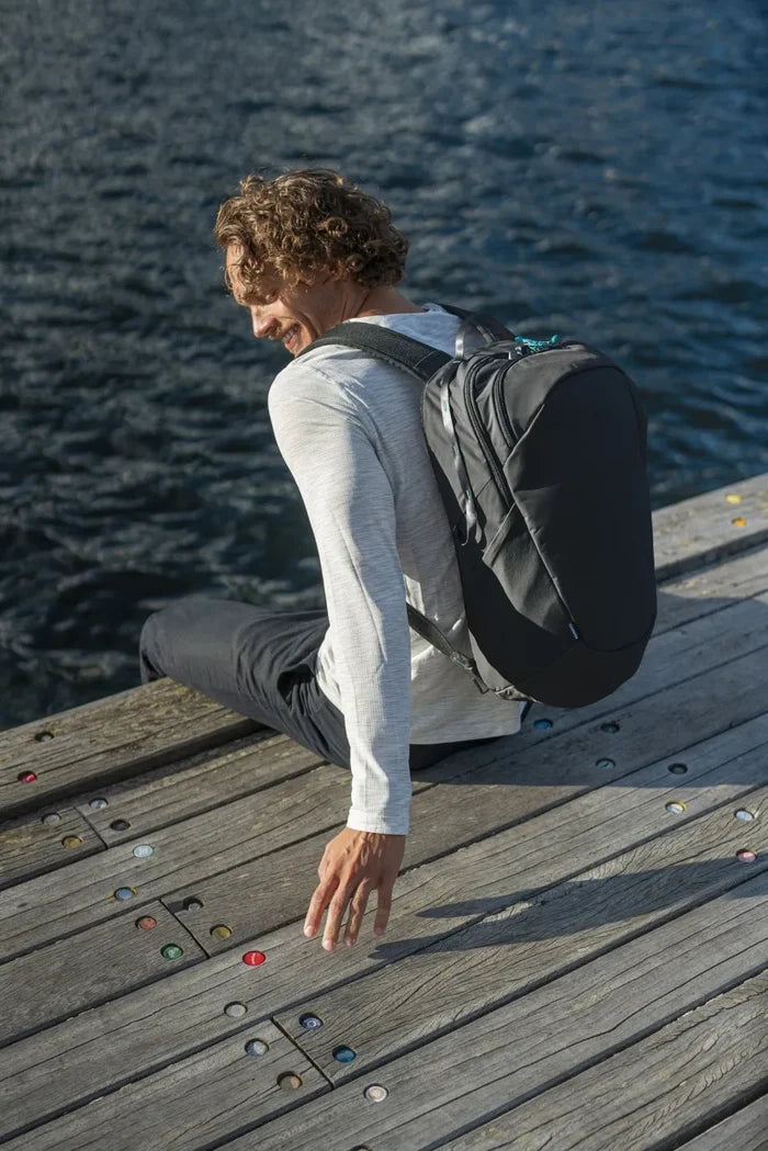 Pacsafe Eco 25L Anti-Theft 16" Laptop Backpack in Black, worn on both shoulders by a man sitting on a boardwalk by the sea