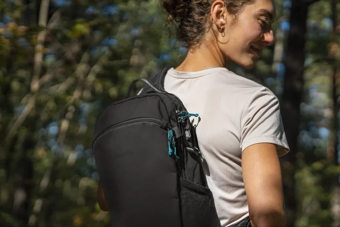 Pacsafe ECO Anti-theft 12L Sling Backpack in Black, worn on both shoulders by a woman walking through woodland