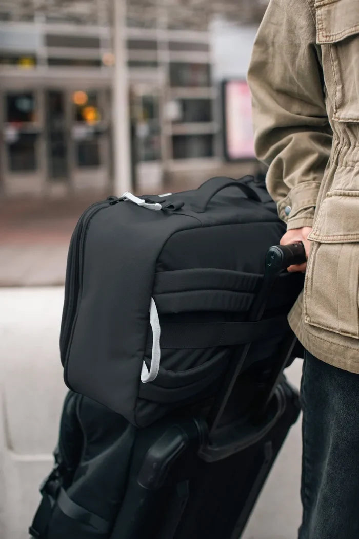 Pacsafe GO 34L Anti-Theft Carry-on Backpack in Jet Black, hooked over the handle of a wheeled case at an airport kerb