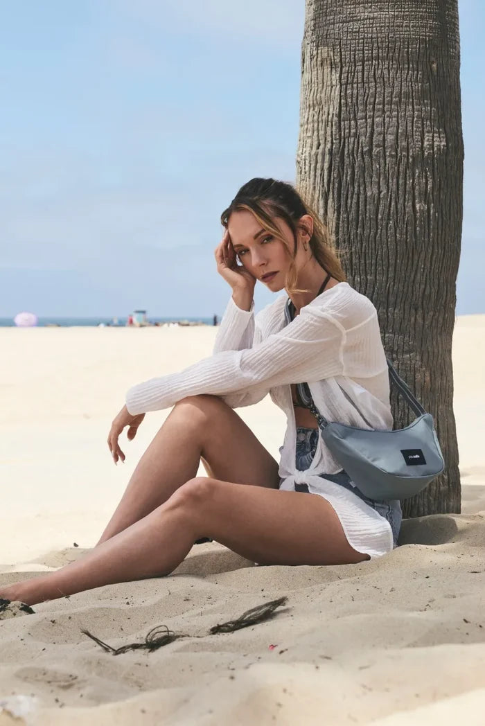 Pacsafe Go Anti-Theft Lunar Crossbody Bag in Fresh Mint, worn at the hip by a woman sitting against a palm tree on the sand