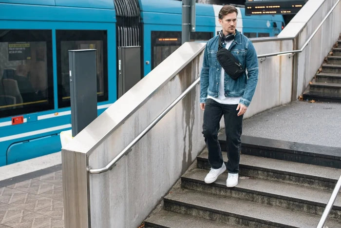 Pacsafe V Action Anti-Theft Sling Pack in Jet Black, worn crossbody by a man walking down station steps past a tram