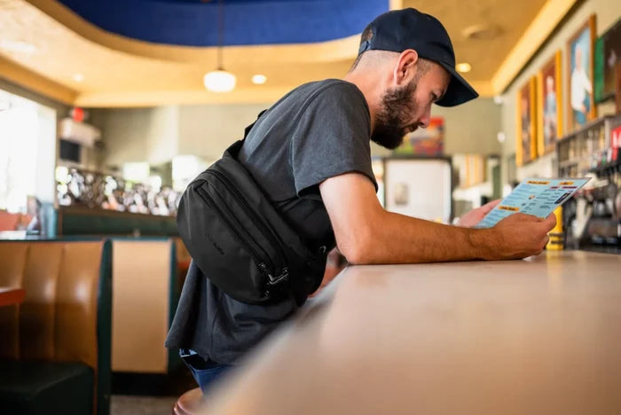 Pacsafe V Anti-Theft Hip Pack in Jet Black, worn crossbody by a man reading a menu at a diner counter