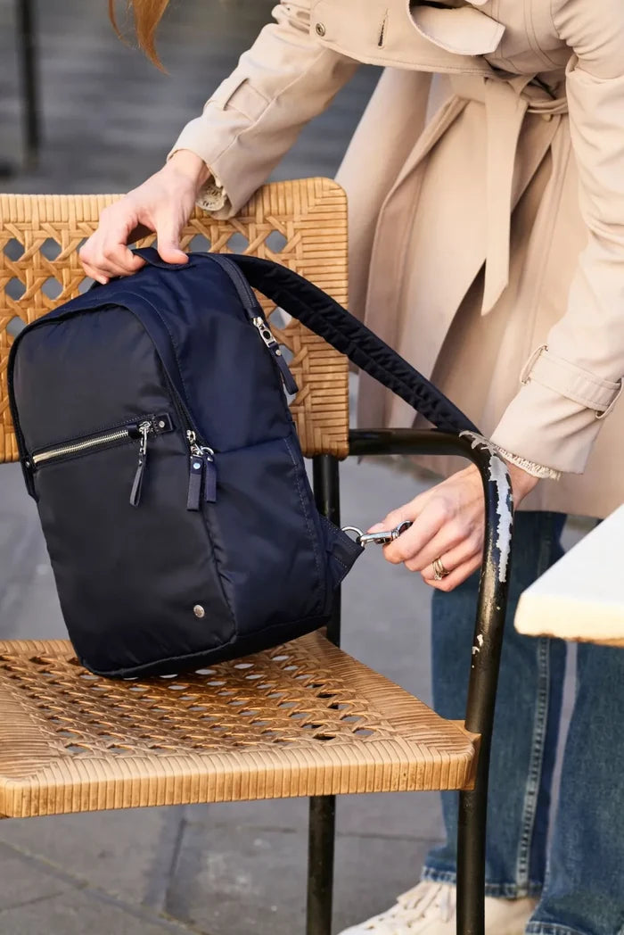 Pacsafe W 10L Anti-theft 13" Laptop Backpack in Navy, lifted from a cane chair by its top handle at a cafe table