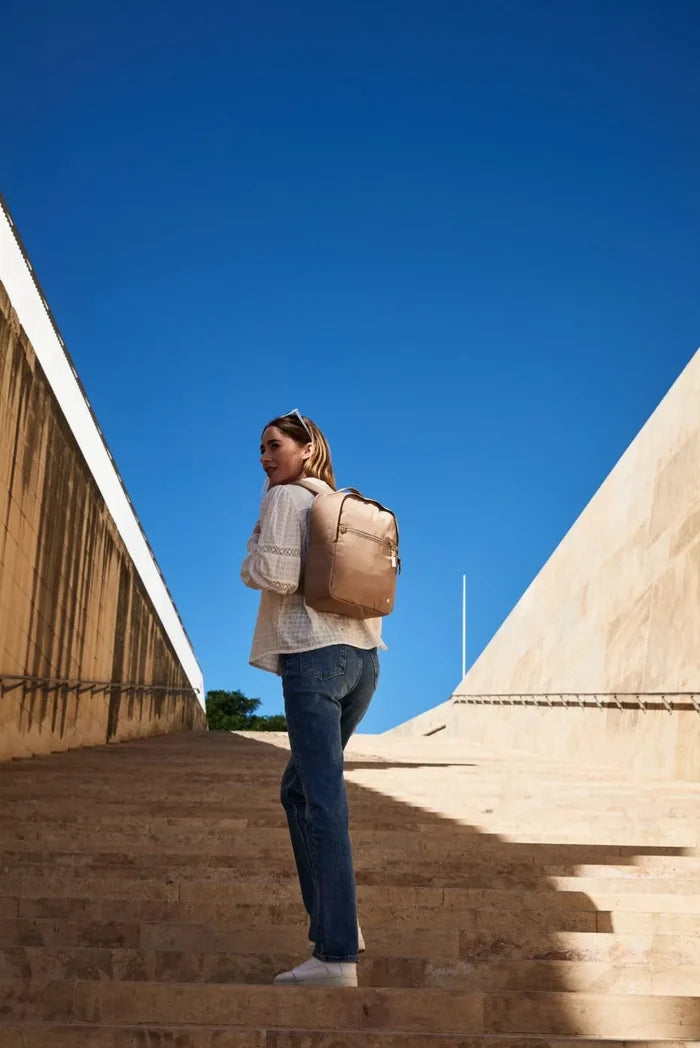 Pacsafe W 10L Anti-theft 13" Laptop Backpack in Taupe, worn on both shoulders by a woman climbing sunlit stone steps